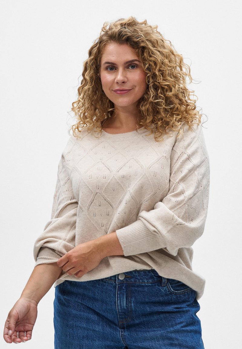 Light beige knitted sweater featuring a diamond pattern, wide sleeves, and ribbed cuffs, paired with blue jeans.