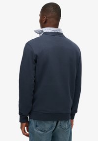 Marineblauwe sweatshirt met een geribde ronde hals, gestructureerde stof en een gelaagde overhemdkraag die aan de achterkant te zien is. Gecombineerd met een blauwe spijkerbroek.