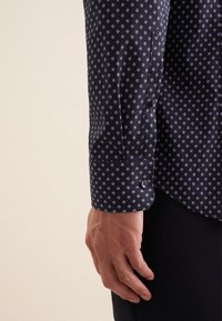 Camicia blu scuro con piccolo motivo a rombi bianchi, maniche lunghe, polsini abbottonati, texture del tessuto liscia. Dettaglio del braccio visibile.