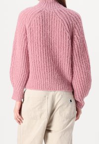 Rosa gestrickter Pullover mit hohem Kragen, gerippter Textur und überschnittenen Schultern. Kombiniert mit hellbeigen Hosen mit Gesäßtaschen.