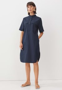 Navyblaues Hemdblusenkleid mit kurzen Ärmeln, Kragen, zwei Fronttaschen, Knopfleiste und seitlichen Schlitzen; aus leichtem Stoff gefertigt.