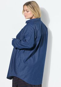 Camicia in denim blu scuro con vestibilità rilassata, maniche lunghe, colletto a bottone e orlo curvo. Presenta una tasca sul petto.