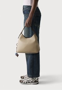 Borsa a tracolla in pelle beige con una silhouette morbida, rifiniture marrone scuro e un accento decorativo in pom-pom bicolore. Indossata con jeans blu oversize e sneakers.