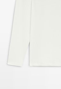 Hvid langærmet beklædning med enkel manchet, ensartet design og diskret sømdetalje på en flad hvid baggrund.