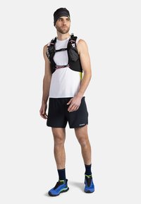 Gilet idratante nero con spalline regolabili, indossato sopra una maglietta bianca senza maniche e pantaloni neri, abbinato a scarpe sportive blu e verdi.