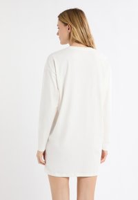 Biała sukienka z długim rękawem o luźnym kroju. Miękka tkanina, okrągły dekolt oraz delikatnie zaokrąglony dół. Brak wyraźnych wzorów i ozdób.