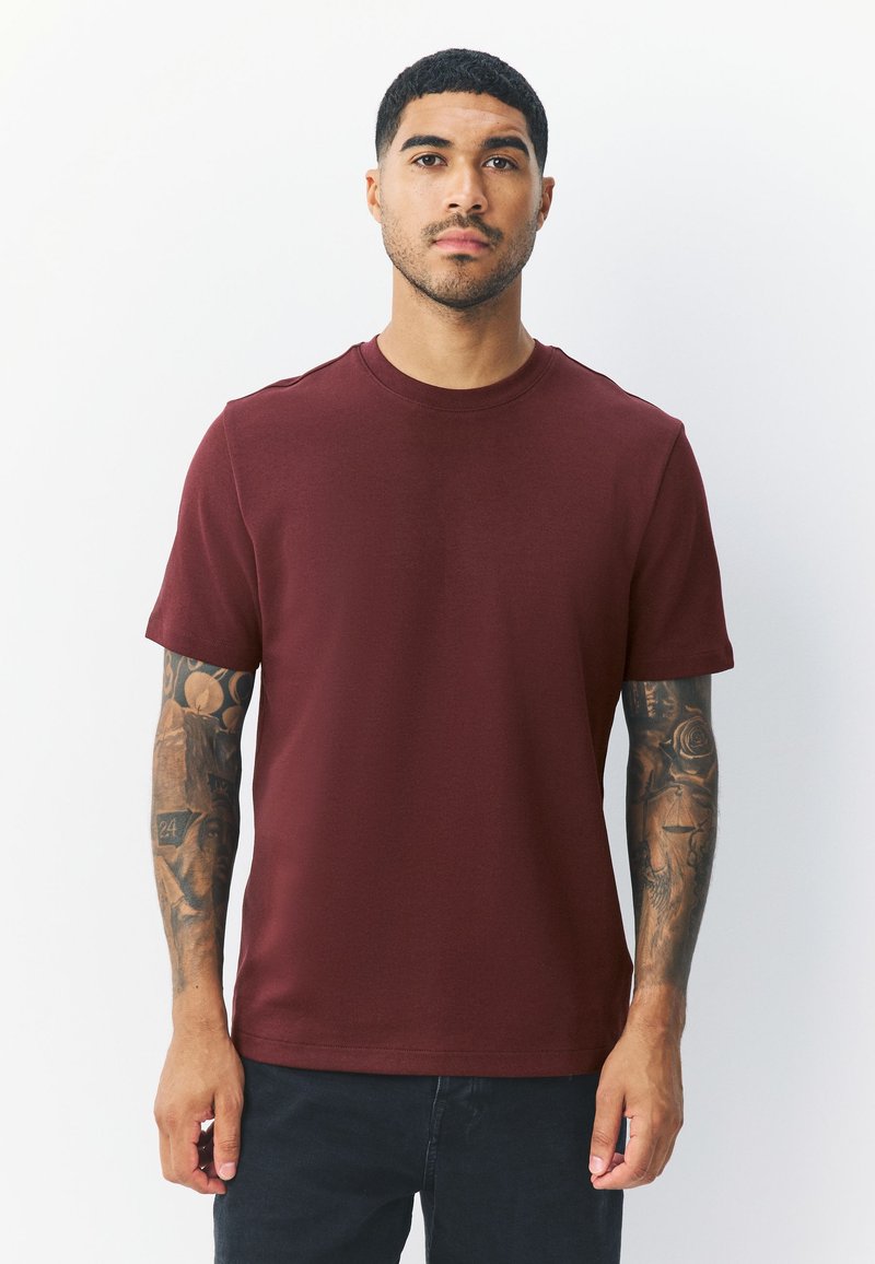 Burgundy short-sleeve t-shirt with a crew neckline, smooth texture, and minimal stitching details, paired with dark pants and tattooed arms.