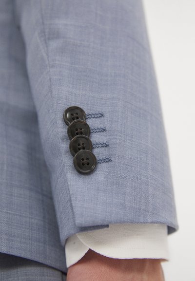 Manica di giacca blu chiaro con quattro bottoni neri, impunture blu a contrasto e polsino di camicia bianca visibile sotto la giacca.