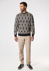 Suéter negro con un patrón geométrico en crema y beige, escote redondo, puños acanalados, combinado con pantalones beige y zapatillas negras.