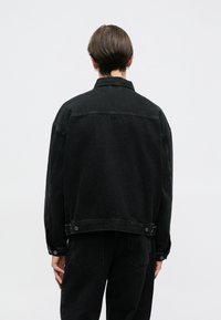 Casaco de ganga preto com corte descontraído, apresentando uma gola, fechos de botões e costuras visíveis. O tecido tem um acabamento texturizado.