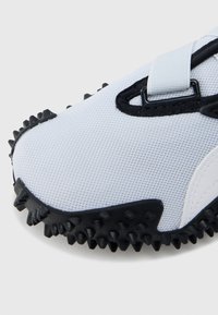 Biały but sportowy z teksturowaną, siateczkową cholewką, czarnymi akcentami oraz kolczastą gumową podeszwą zapewniającą przyczepność. Zapięcie na pasek.