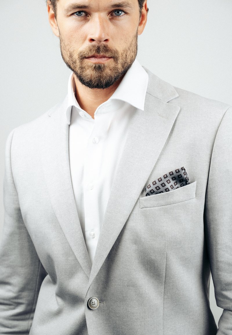 Mężczyzna z jasnym zarostem, ubrany w jasnoszary marynarkę, białą koszulę oraz wzorzysty poszetkę na tle jednolitego tła.
