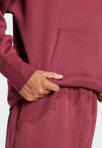 Felpa bordò con tasca e polsini a costine, caratterizzata da una texture morbida e una vestibilità rilassata. Mano posizionata all'apertura della tasca.