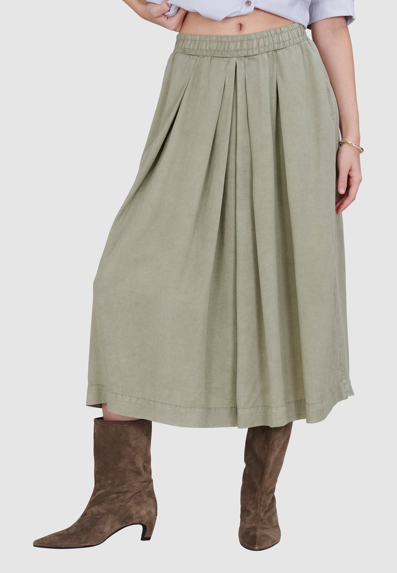 Knie-lange, geplooide rok van licht olijfgroene stof met een elastische tailleband. Gecombineerd met bruin suède enkellaarsjes met een spitse neus.