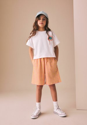 Fille debout, les mains dans les poches, portant un t-shirt blanc avec une fleur, un short à carreaux rose et jaune, une casquette bleu clair, des chaussettes blanches et des baskets.