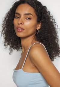 Top sem mangas azul claro com decote quadrado, feito de um tecido suave. A modelo tem cabelo encaracolado e usa brincos de argola. Makeup natural.