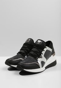 Un par de zapatillas deportivas negras y plateadas metálicas con medias suelas blancas y cordones negros sobre una superficie blanca.