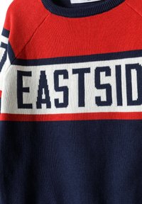 Pull en maille rouge, bleu marine et blanc avec "EASTSIDE" en grandes lettres sur le devant. Il a un col rond et des poignets côtelés.