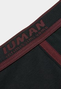 Tejido negro con una cinturilla elástica burdeos que presenta el texto en relieve "IUMAN". Textura suave con detalles de costura en contraste.