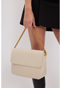 Beige neplederen handtas met een klepsluiting, voorzien van een gouden schouderband en subtiele stikdetails langs de randen.