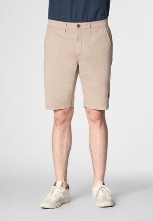Pantaloni corti beige con design a vita piatta, passanti per cintura, due tasche laterali e un discreto dettaglio del logo sull'orlo, indossati con sneakers bianche.