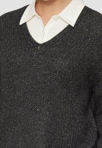 Koksgrå striktrøje med V-hals og synlig struktur. Båret over en hvid skjorte med krave, med fine metalliske tråde gennemgående.