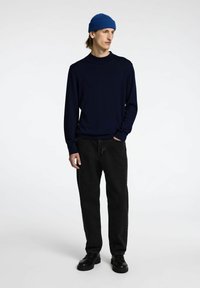 Maglione a maniche lunghe blu scuro, jeans neri e scarpe nere. Un berretto lavorato a coste blu. Il modello è in piedi con le mani nelle tasche contro uno sfondo chiaro.