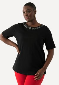 Camiseta negra de manga corta con cuello redondo adornado con tachuelas plateadas, que presenta un ajuste relajado y un material de algodón suave.