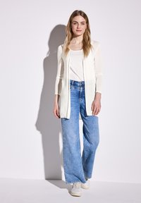 Cardigan créme léger et long sur un haut blanc, associé à un jean en denim bleu clair ample et des baskets blanches. Texture lisse, design décontracté.