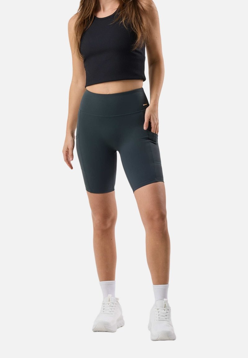 Γυναίκα που φοράει μαύρο αμάνικο crop top, σκούρο γκρι ποδηλατικά σορτς με πλαϊνή τσέπη, λευκές κάλτσες και λευκά αθλητικά παπούτσια, στέκεται σε λευκό φόντο.