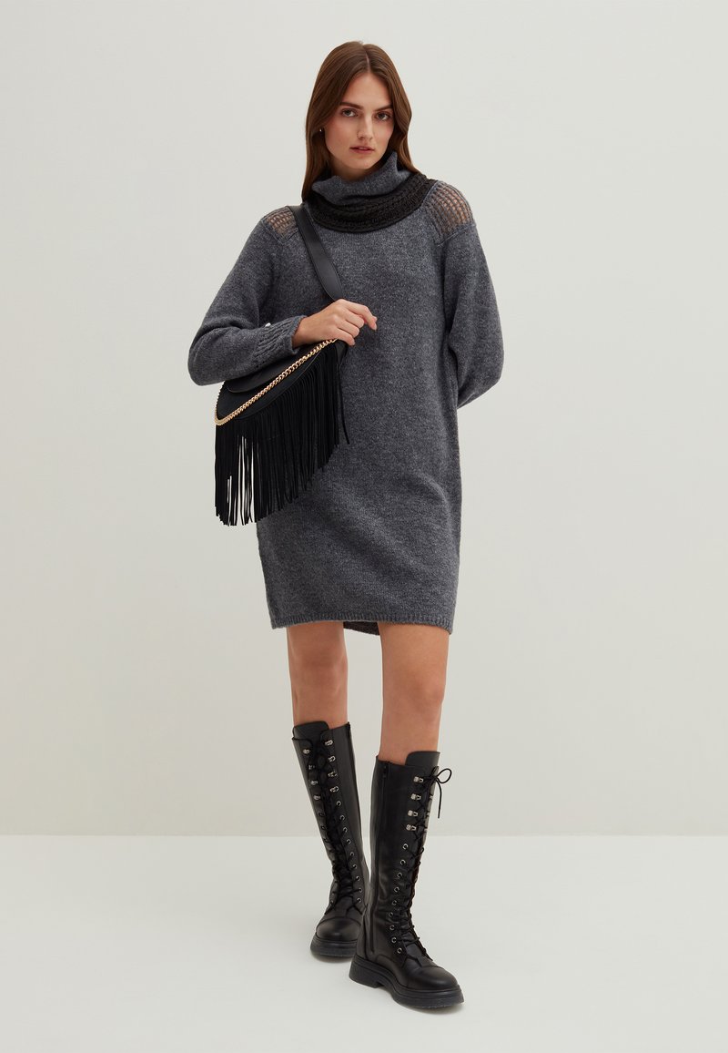Abito lavorato a maglia grigio con colletto alto e dettaglio delle spalle in maglia aperta, abbinato a stivali neri alti fino al ginocchio con lacci e una borsa nera con frange.