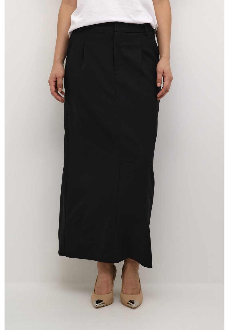 Jupe noire mi-longue en tissu lisse avec une coupe ajustée. Elle possède des poches avant et un design de coutures épurées, associée à des talons beiges.