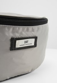 Szara torba z nylonu o zaokrąglonym kształcie, z czarnym zamkiem błyskawicznym oraz prostokątną czarną metką z białym tekstem.