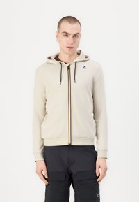 Beige cipzáras kapucnis pulóver, húzózsinórral, elől színes csíkkal és logóval a mellkason. Texturált anyag.