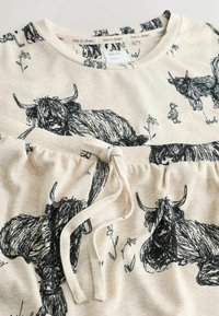 Set de pijamale bej cu imprimeuri negre în stil schiță cu bovine Highland și accente florale. Bluza are guler rotund; pantalonii au șnur ajustabil.