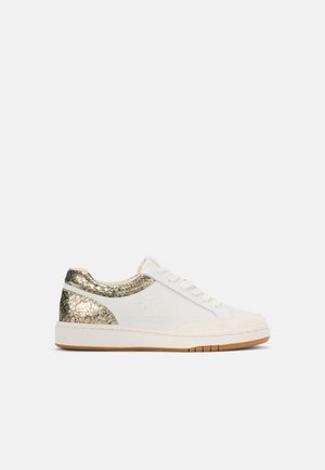 Fehér sneaker arany színű, textúrázott díszítésekkel a sarokon és az oldalakon. Lapos gumitalppal, kerek orral és fűzős elemmel rendelkezik.