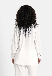 Hvid, tekstureret bluse med lange ærmer, buet kant og knappede manchetter, båret med matchende bukser, set fra ryggen.
