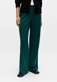 High-waisted, wide-leg trousers in deep teal fabric. Smooth texture with a slight sheen. Paired with a black zip-up jacket and flat shoes.