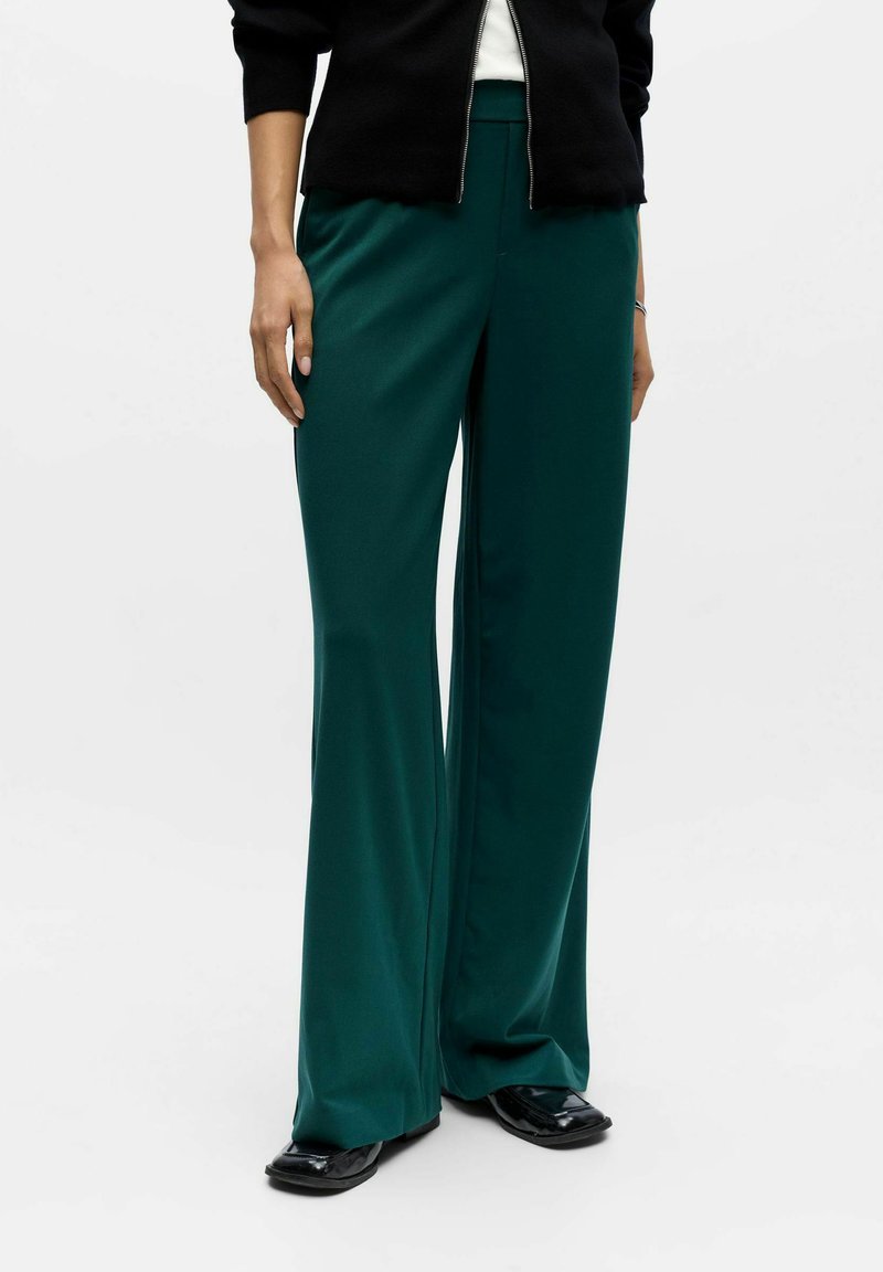 High-waisted, wide-leg trousers in deep teal fabric. Smooth texture with a slight sheen. Paired with a black zip-up jacket and flat shoes.