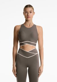 Žena nosi bezrukavi taupe crop top i tajice visokog struka s kontrastnim svjetlim obrubima i izrezima na torzu.