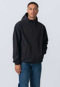 Sort letvægtsjakke med frontlynlås, justerbar hætte og elastiske manchetter. Båret over blå jeans ser stoffet glat og mat ud.