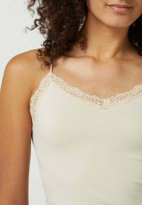Beige top met verfijnde kantafwerking langs de halslijn en verstelbare bandjes, met een gladde, strakke textuur.