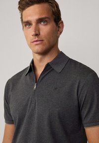 Polo shirt grigio scuro realizzato in tessuto liscio, con una placca a mezza zip e un logo discreto sul petto. Maniche corte e colletto classico.
