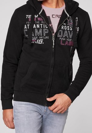 Mężczyzna w czarnej bluzie z suwakiem, z białym i różowym napisem, pod którą ma różową koszulkę, oraz w jasnoniebieskich dżinsach, dłonią poprawiający zamek błyskawiczny.