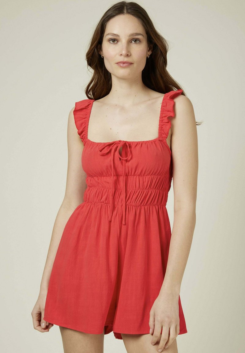 Robe d'été rouge avec un décolleté carré, des bretelles à volants et un lien ajustable à l'avant. Présente une taille smockée et un short ample.