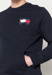 Marineblauwe sweatshirt met ronde hals, voorzien van een geborduurd meerkleurig vlagontwerp op de borst en een voorzak.