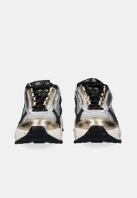 Par de zapatillas deportivas negras, plateadas y doradas con cordones negros, mediasoles blancos y suelas texturizadas negras sobre un fondo blanco.