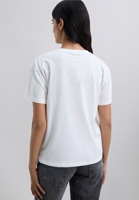 Donna con lunghi capelli scuri rivolta di spalle, che indossa una semplice maglietta bianca a maniche corte e jeans neri sbiaditi, su uno sfondo chiaro.