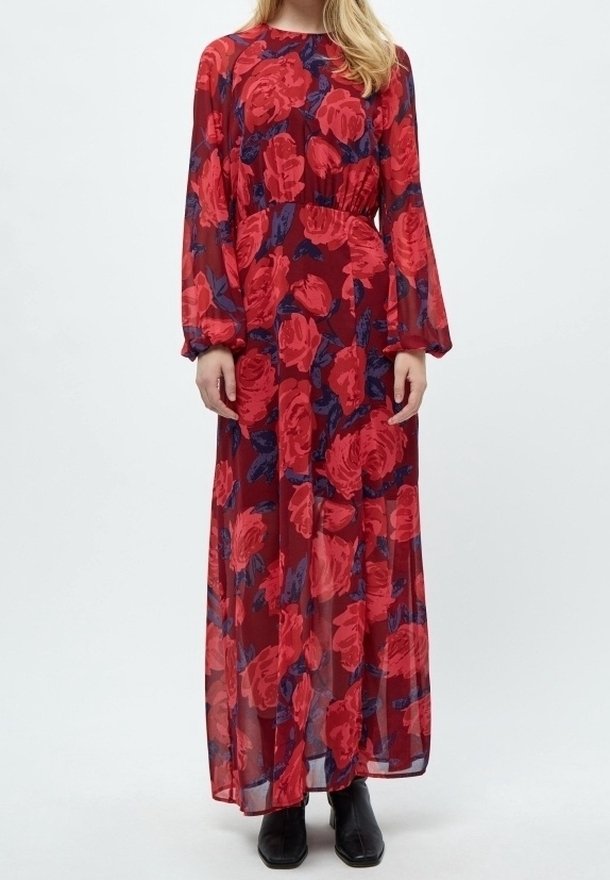 Femme portant une longue robe bordeaux transparente avec de grands motifs floraux rouges et des bottines noires, debout devant un fond uni.