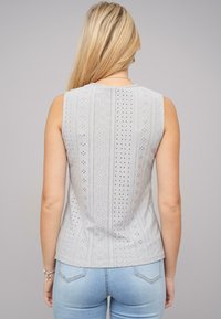 Top gri fără mâneci, cu modele intricate tăiate și un design ajustat, asociat cu blugi albaștri deschis. Detalii texturate ale materialului vizibile.